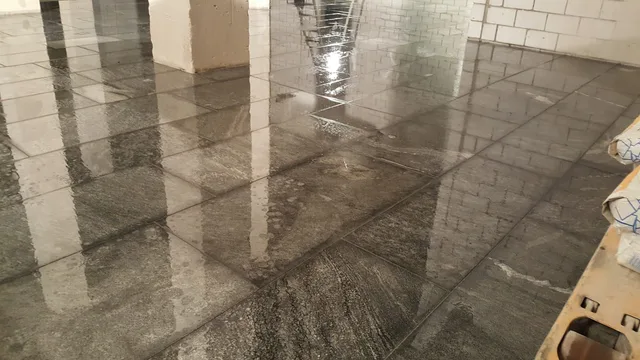 Weite Ansicht eines nassen, dunklen Plattenbodens im modularen Rechteckverband zwischen Wänden und mehreren Stützen.
