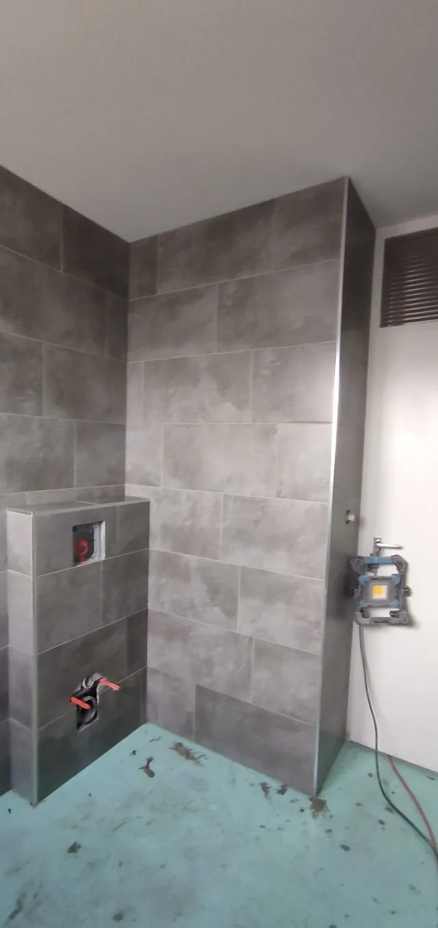 Graues Bad im Ausbau mit raumhoher Wandbekleidung, verfliester WC-Vorwand, offenen Anschlussstellen und geschütztem Rohboden.