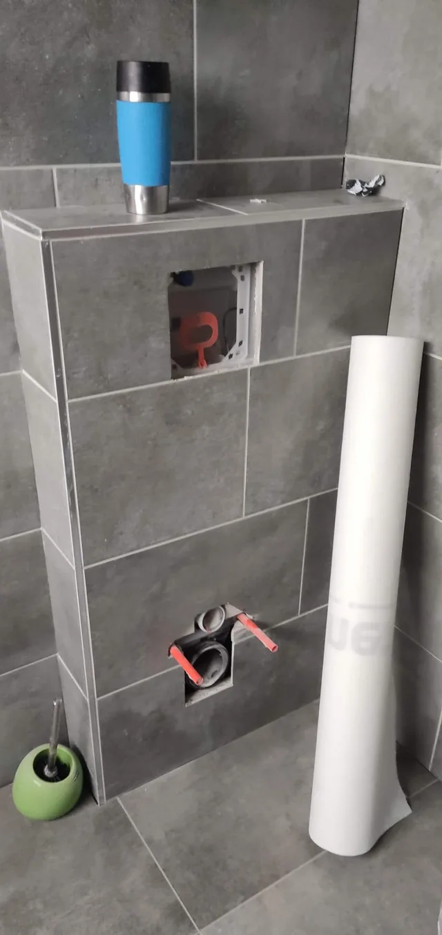 Nahansicht der grau verfliesten WC-Vorwand mit offenen Aussparungen für Betätigung und Anschlüsse sowie belegtem Boden.