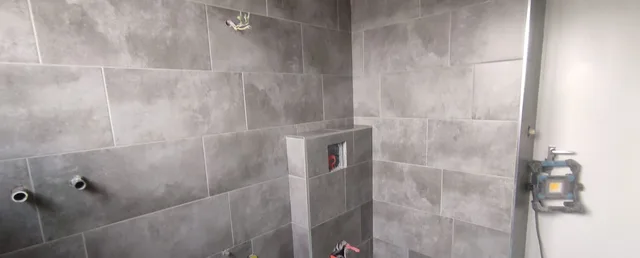 Breite Ansicht des grauen Bades im Ausbau mit WC-Vorwand, Wandbekleidung über mehrere Ebenen und offenen Leitungsanschlüssen.
