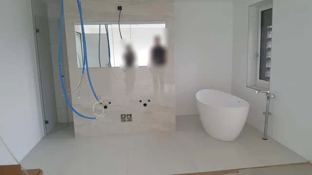 Breite Ansicht des hellen Badezimmers im Ausbau mit freistehender Wanne, Glasdusche, grossformatigen Platten und offener Waschtischzone.