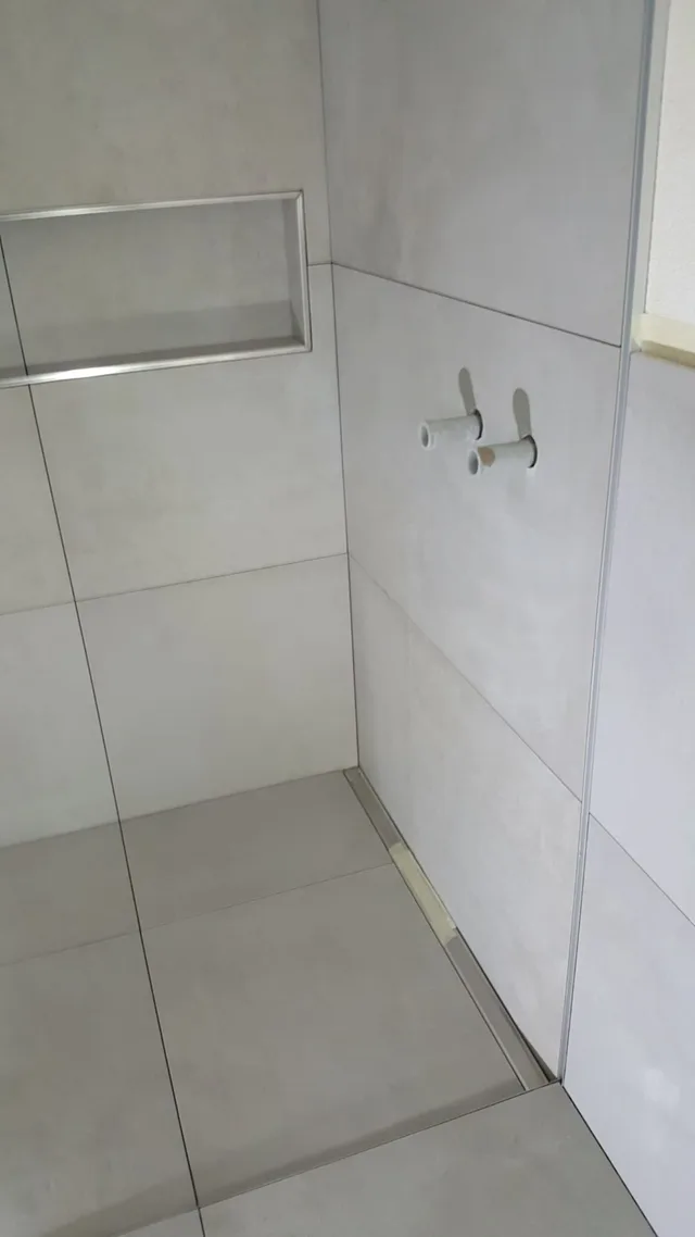 Seitlicher Detailblick in die hellgraue Dusche mit grossformatigen Platten, Wandnische, zwei Rohranschlüssen und Linienablauf.