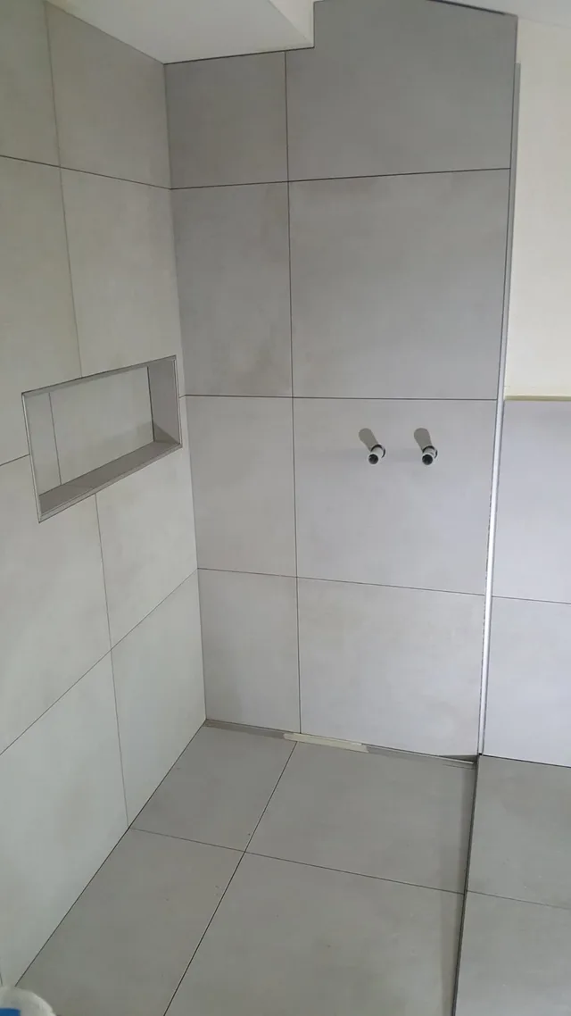 Frontale Gesamtansicht der hellgrauen Dusche im Ausbau mit Wandnische, offenen Armaturenanschlüssen und Linienablauf.