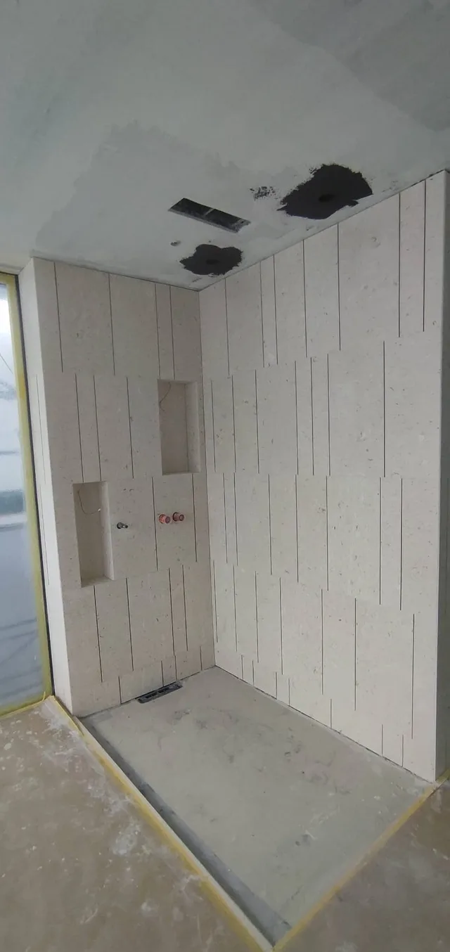 Dusche im Ausbau mit schmalen, hochkant angeordneten hellen Platten, zwei Wandnischen und bodennaher Entwässerung.