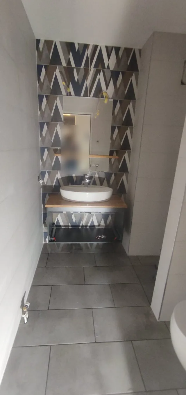 Frontaler Blick auf die geometrische Akzentwand mit Spiegel, Holzablage und weissem Aufsatzbecken im kleinen WC-Raum.